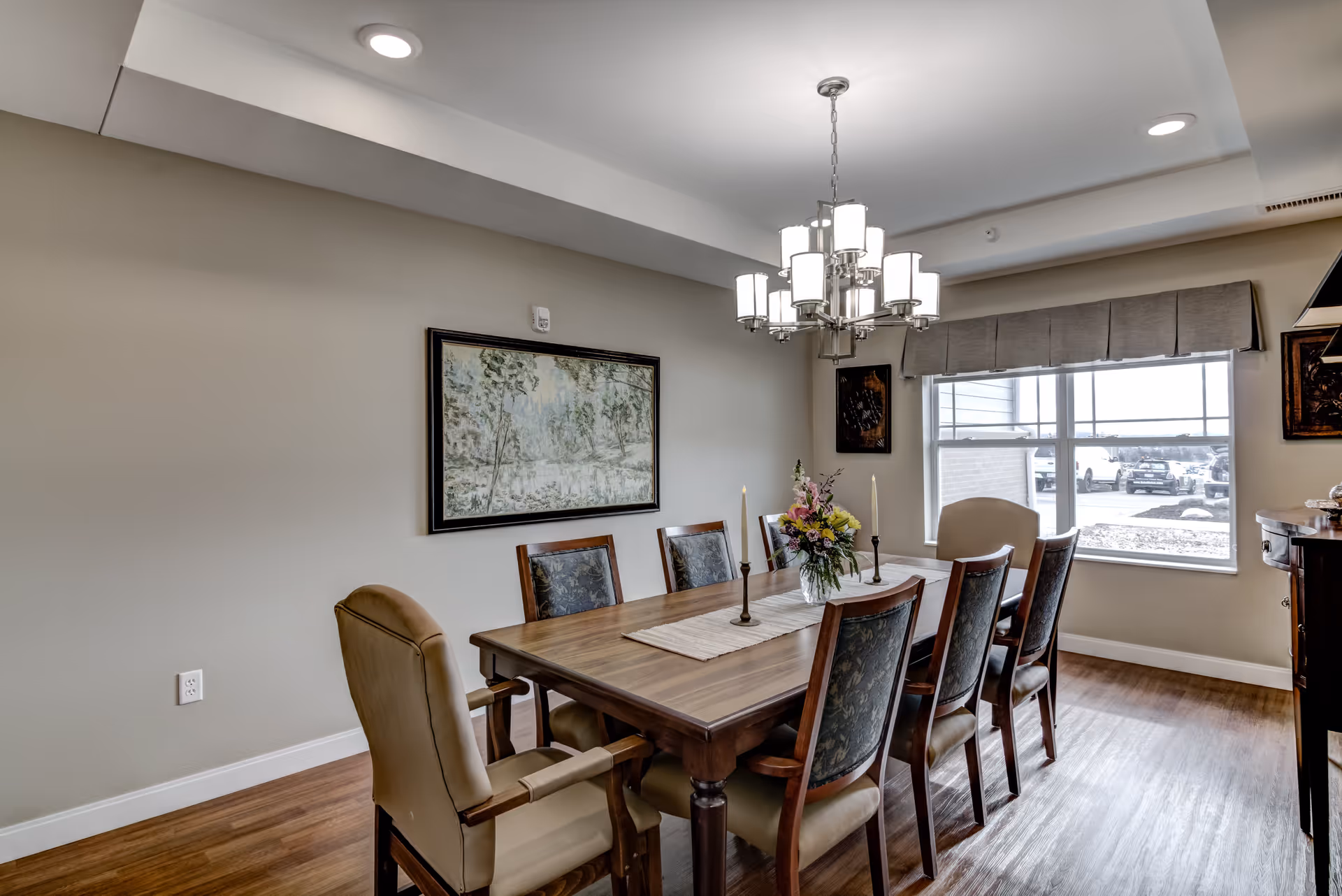 A dining room with a wooden table surrounded by eight chairs, two of which have armrests and beige upholstery. The table is decorated with a floral centerpiece and two tall candles. A modern chandelier with multiple lights hangs above the table. There is a large window with a gray valance letting in natural light, and framed artwork is displayed on the beige walls. The floor is wooden.
