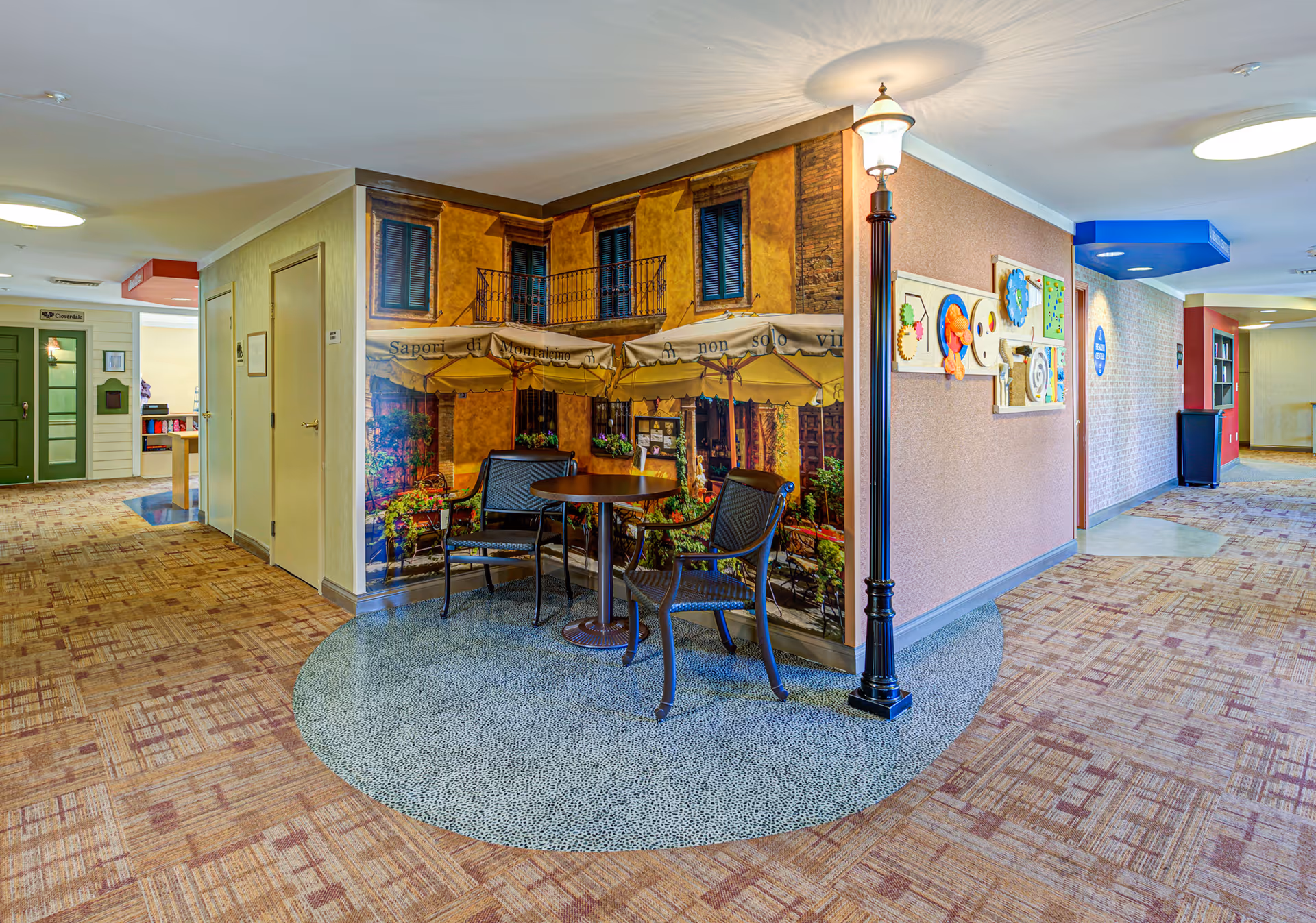 A hallway in a senior living facility with carpeted floors and beige walls. There is a small seating area with two chairs and a round table placed on a circular patterned rug. Behind the seating area is a wall mural depicting an outdoor café scene with umbrellas and a balcony. A decorative street lamp stands next to the seating area. The hallway extends in both directions with doors and wall-mounted decorations visible.