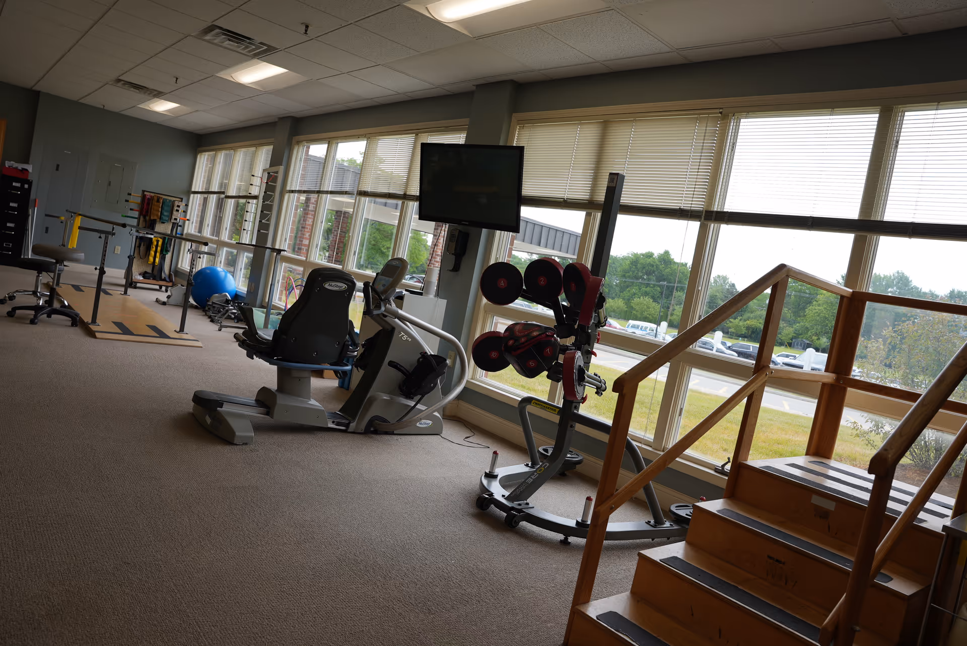 A rehabilitation or physical therapy room with exercise equipment including a recumbent bike, parallel bars, a blue exercise ball, and a wooden staircase for mobility training. Large windows provide natural light and a view of the outside parking area and greenery.