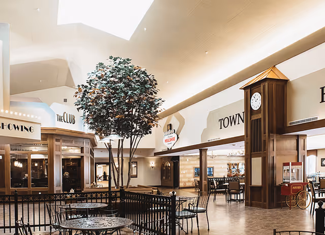 Interior view of a senior living facility common area with a tall artificial tree in the center, metal tables and chairs, a large wooden clock tower, and various storefront-style signs including 'The Club' and 'Town'. The space has high ceilings and warm lighting.