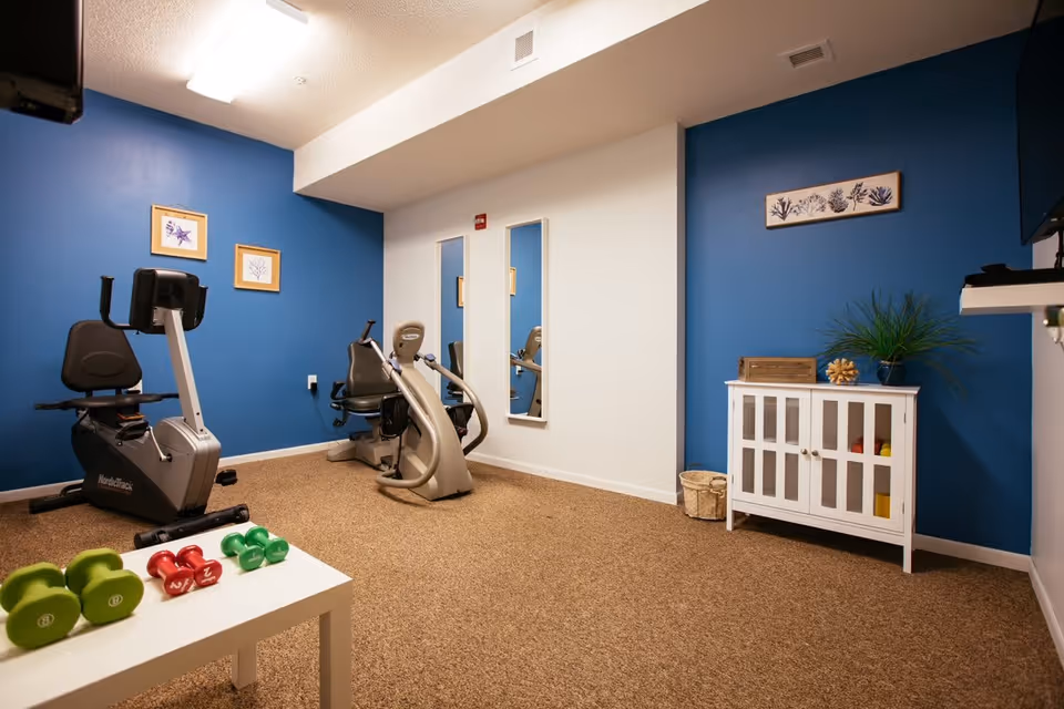 A small fitness room with blue and white walls, featuring two exercise bikes, a white table with green and red dumbbells, a white cabinet with glass doors, a plant on top, and framed artwork on the walls.
