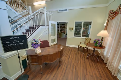 Interior view of a senior living facility reception area with a wooden desk, a sign that reads 'Welcome to The Manor at Victoria Park,' a staircase with white railings, a small round table with a lamp and flowers, and a chair. There is a window into another room with bookshelves and a curtain on the right side.