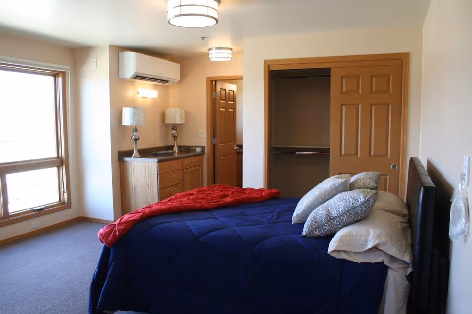 A bedroom with a large window on the left, a bed with a blue comforter and multiple pillows, a red blanket draped over the foot of the bed, a wooden dresser with two lamps, and an open closet with sliding wooden doors.