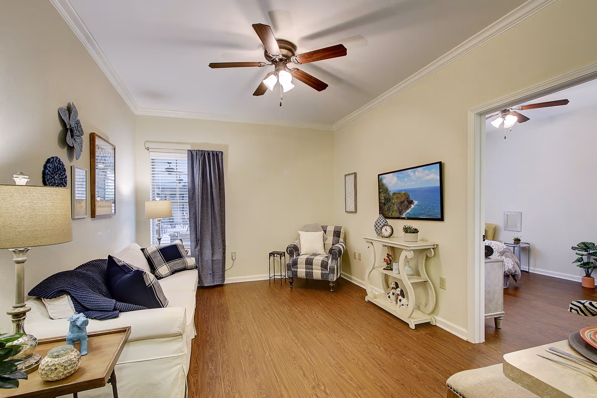 A cozy living room with a white sofa adorned with dark blue and plaid pillows, a floor lamp, and wall decorations. There is a ceiling fan with lights above, a window with a curtain, a patterned armchair, and a white console table with decorative items and a wall-mounted TV showing a coastal scene. The room has wooden flooring and light-colored walls. An adjacent room with a bed and plants is partially visible through an open doorway.