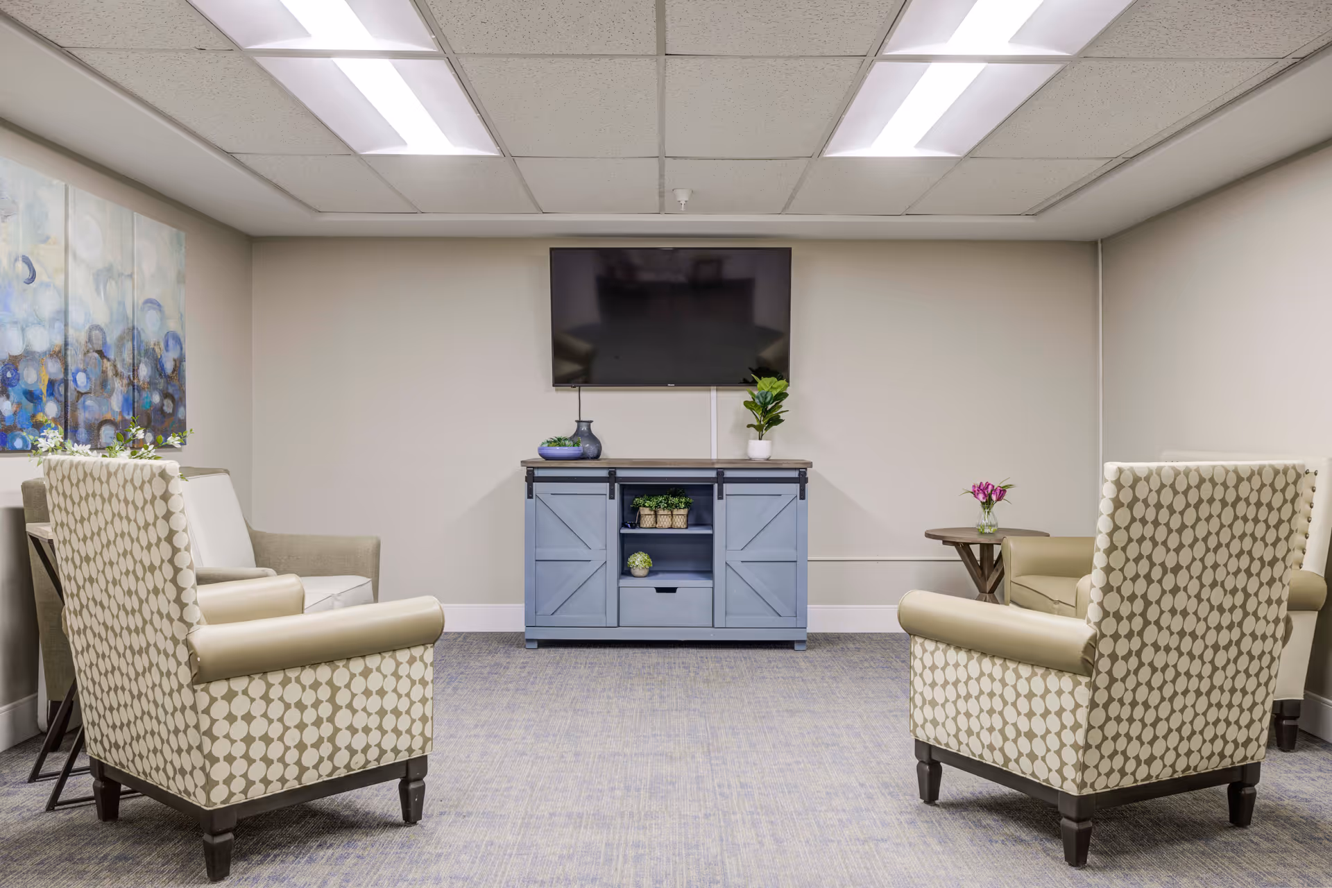 Small seating area with upholstered armchairs facing a wall-mounted TV above a blue cabinet in a bright lounge.