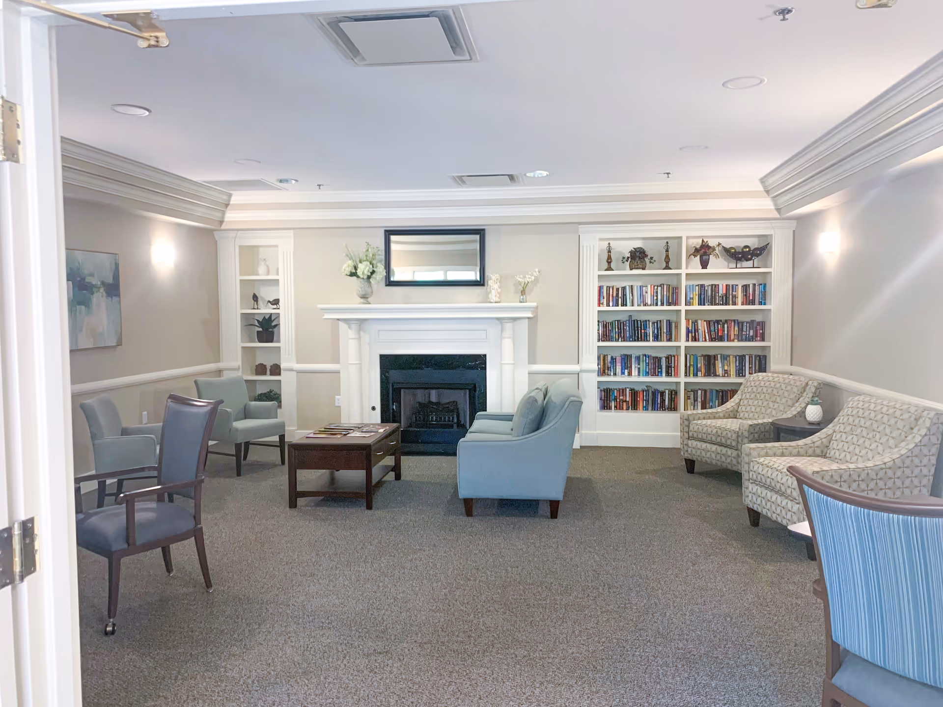A bright common lounge with sofas and chairs arranged around a central fireplace and built-in bookshelves.