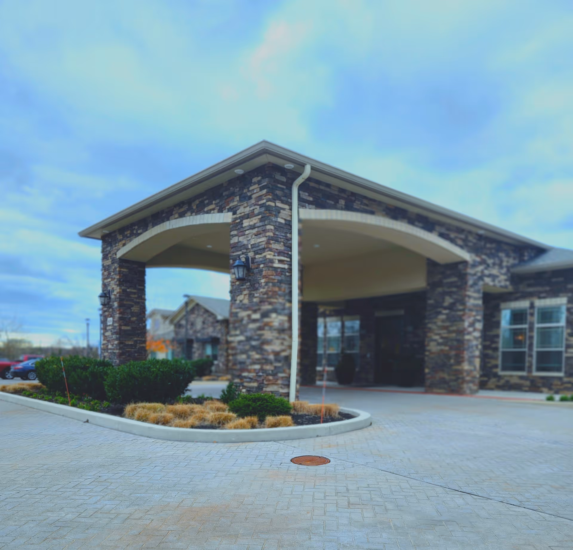 Exterior view of a building entrance with stone pillars and an arched roof covering the driveway. There are some bushes and landscaping in front, and a few parked cars are visible in the background under a cloudy sky.