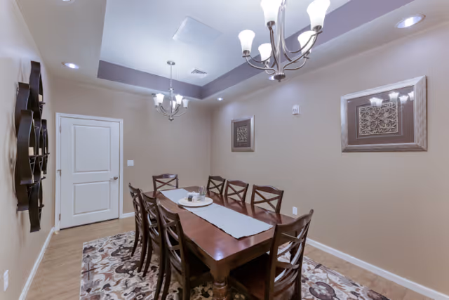 A dining room with a long wooden table surrounded by eight matching wooden chairs. The table has a light gray runner and a decorative centerpiece. The room features beige walls, two framed artworks, a patterned area rug, and two ceiling light fixtures with multiple bulbs.