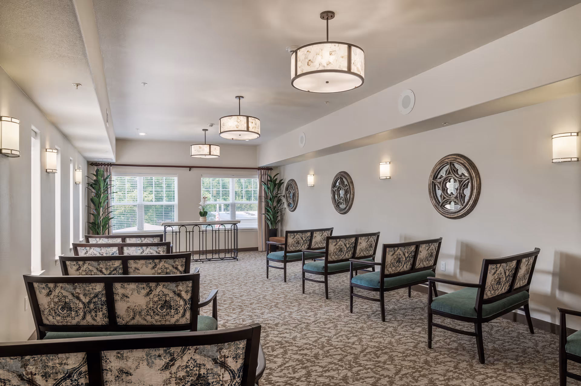 A well-lit room with patterned carpet and multiple chairs arranged in rows facing a window with white blinds. The walls are decorated with round ornamental mirrors and wall sconces. Three ceiling lights hang from the ceiling, and there are two tall potted plants near the window.