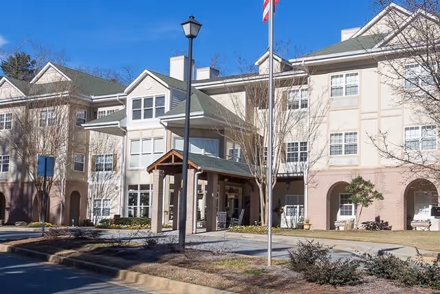 Exterior view of a multi-story senior living facility building with beige and brick facade, a covered entrance with rocking chairs, a flagpole with an American flag, and a streetlamp in front. Trees without leaves and some landscaping are visible around the building.