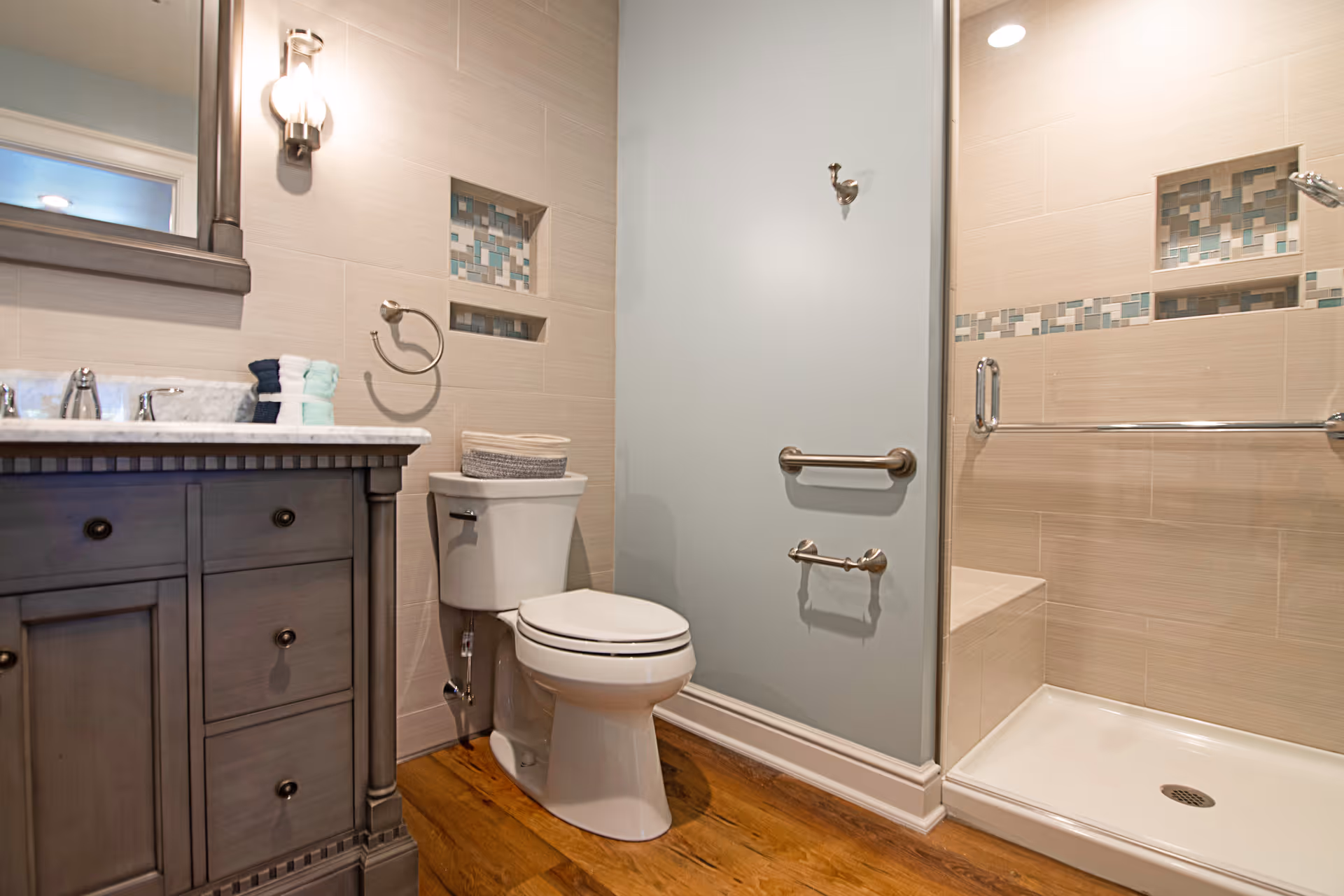 A modern bathroom featuring a wooden vanity with a marble countertop and a mirror above it, a toilet with a basket on top, and a walk-in shower with glass doors and decorative tile accents. The floor is wooden, and the walls are tiled in neutral tones with a light blue accent wall.