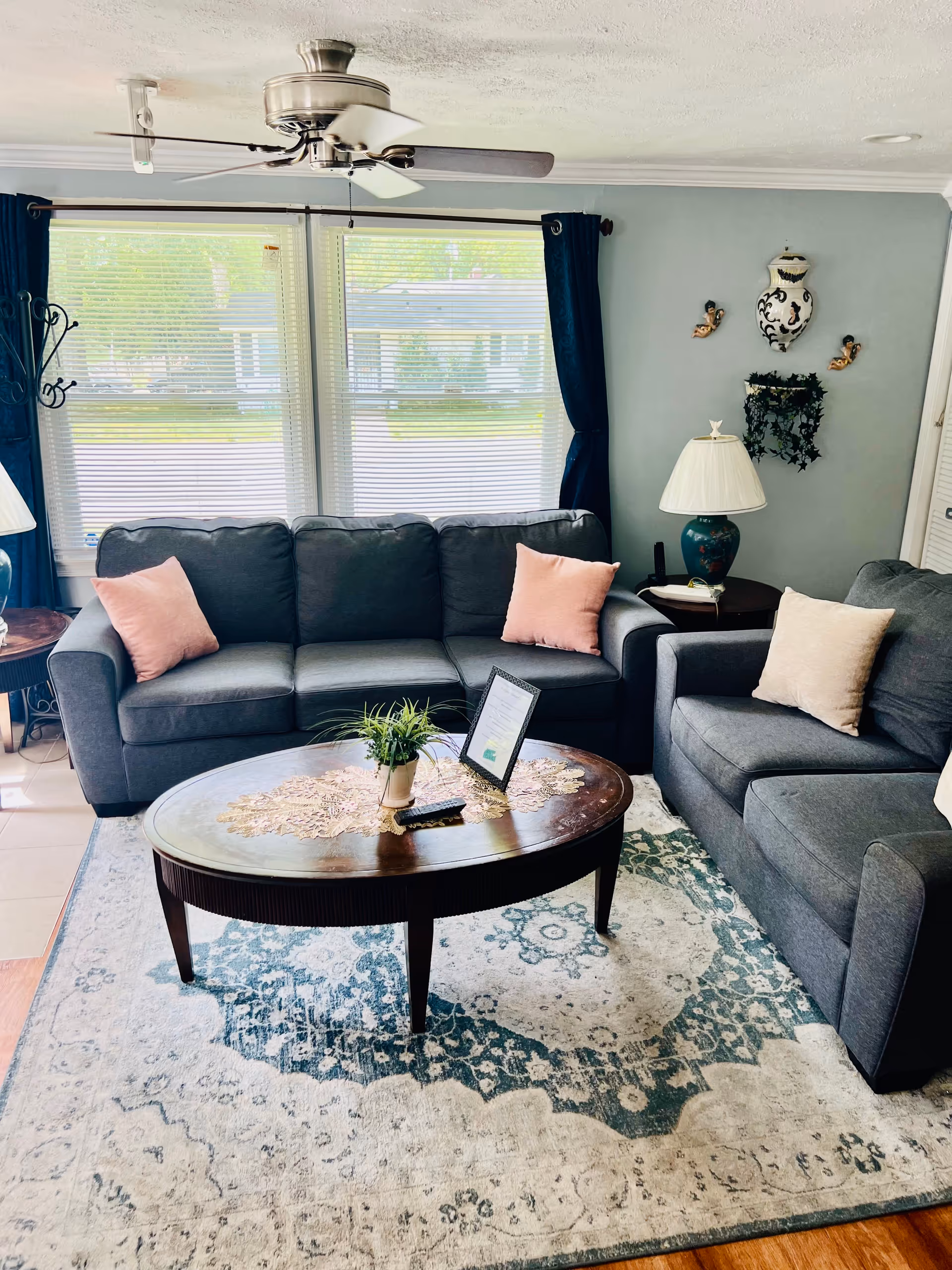 Bright living room with gray sofa and armchair arranged around a wooden oval coffee table on a patterned rug, with large windows, dark curtains and a ceiling fan.