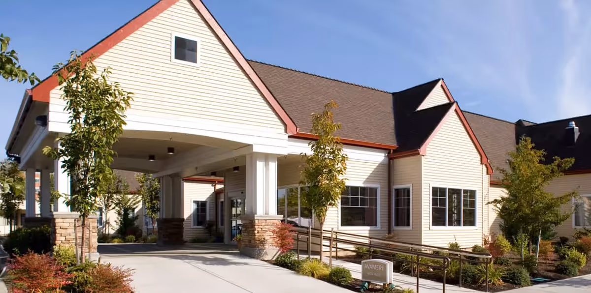 Exterior view of Avamere Rehabilitation of Hillsboro building with beige siding, red trim, and a covered entrance. There are small trees and landscaped bushes around the entrance and a clear blue sky above.