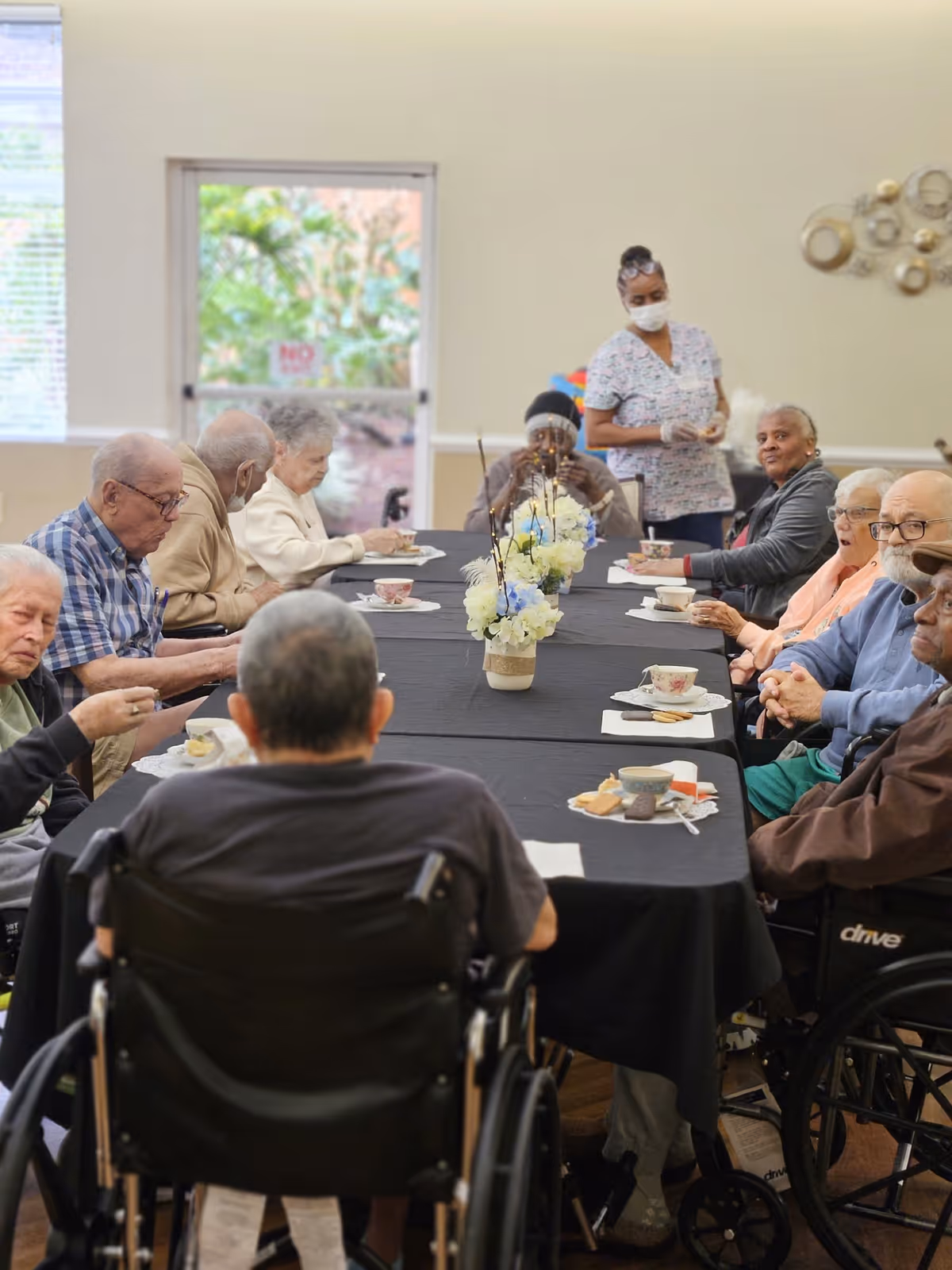 A group of elderly people sitting around a long table covered with a black tablecloth, enjoying tea and snacks. A caregiver wearing a mask and gloves stands nearby. The room has light-colored walls and a window with a view of greenery outside. There are floral centerpieces on the table.