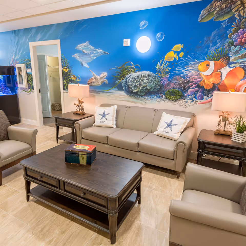 A cozy living room with beige leather sofa and armchairs, a dark wooden coffee table, and side tables with lamps. The wall behind the sofa features a colorful underwater mural with dolphins, a sea turtle, clownfish, and coral. There is a doorway leading to another room and an aquarium visible on the left side.
