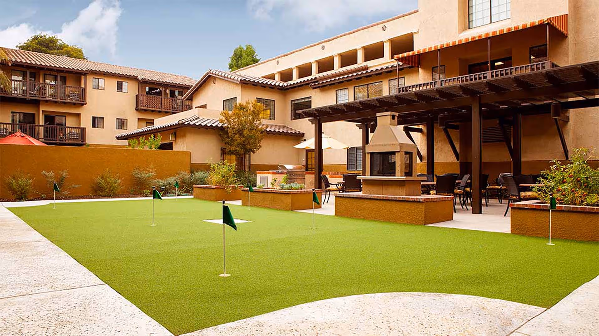 Outdoor courtyard area at Atria Del Rey featuring a putting green with several small flags, surrounded by a beige building with balconies and a covered patio area with tables, chairs, umbrellas, and an outdoor fireplace.