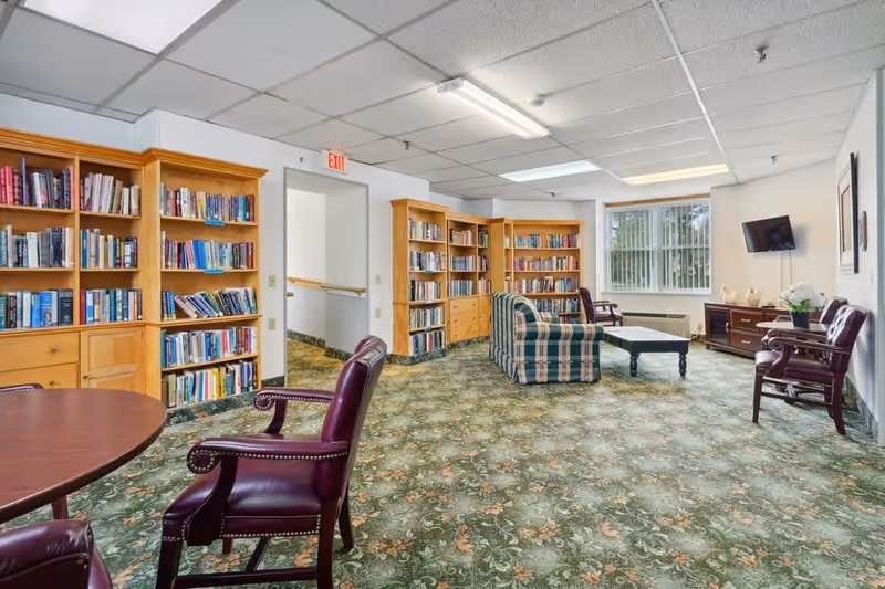 A cozy senior living common room with floral patterned carpet, wooden bookshelves filled with books, a plaid upholstered sofa, several wooden chairs with leather cushions, a coffee table, a wall-mounted TV, and a window with vertical blinds letting in natural light.