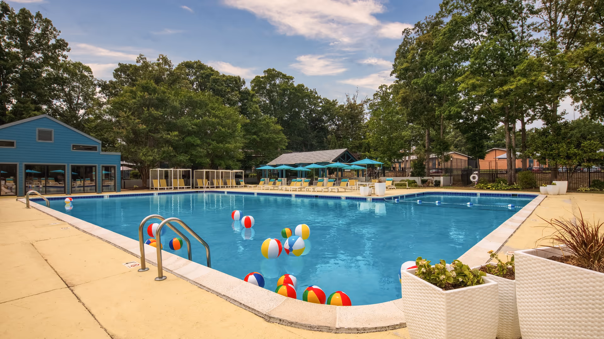 Outdoor swimming pool with colorful beach balls floating on the water, surrounded by a concrete deck with lounge chairs, umbrellas, and large planters. Trees and a blue building are visible in the background under a partly cloudy sky.
