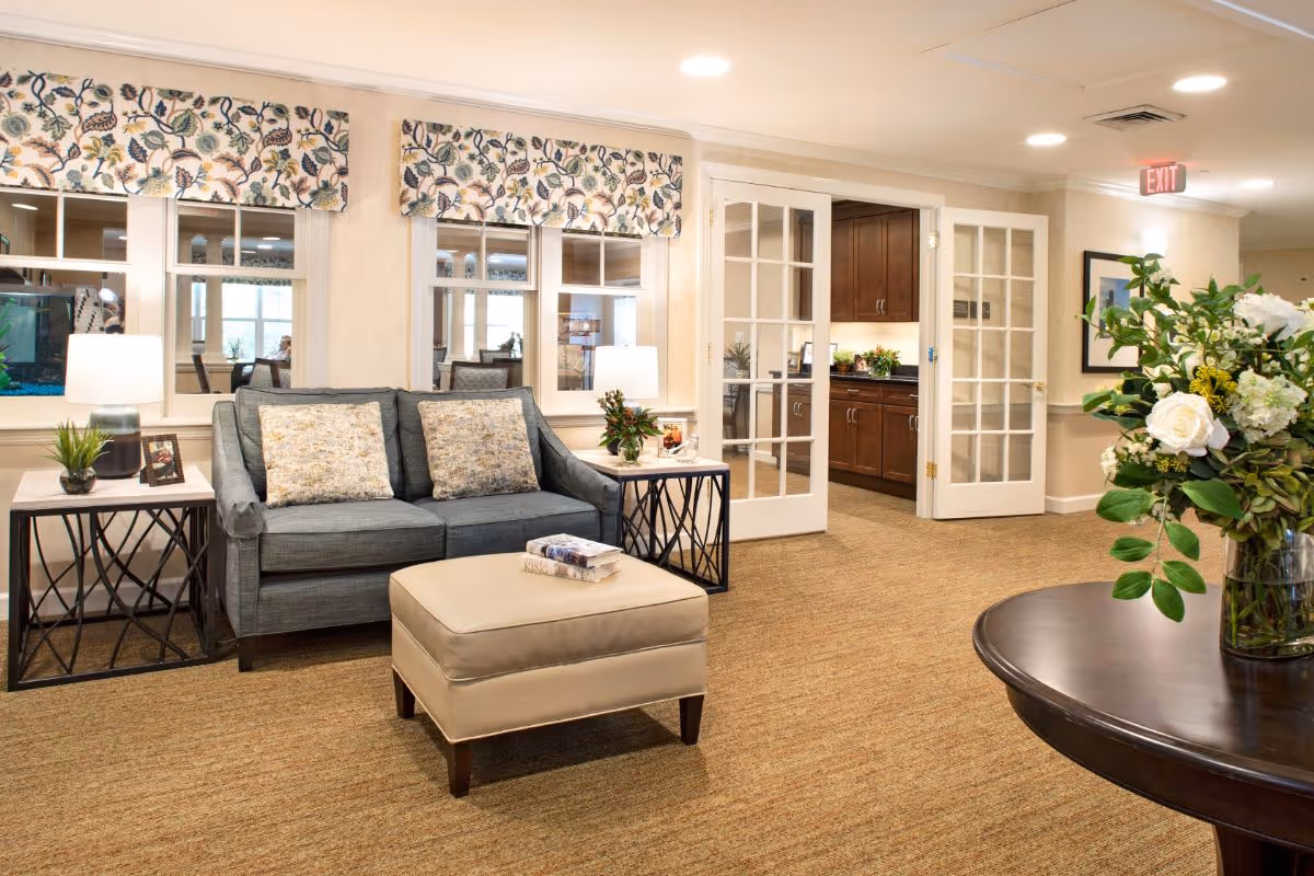A bright, cozy sitting area with a sofa, ottoman, side tables, floral window valances, glass-paned doors to a kitchenette, and a round table with a vase of flowers.