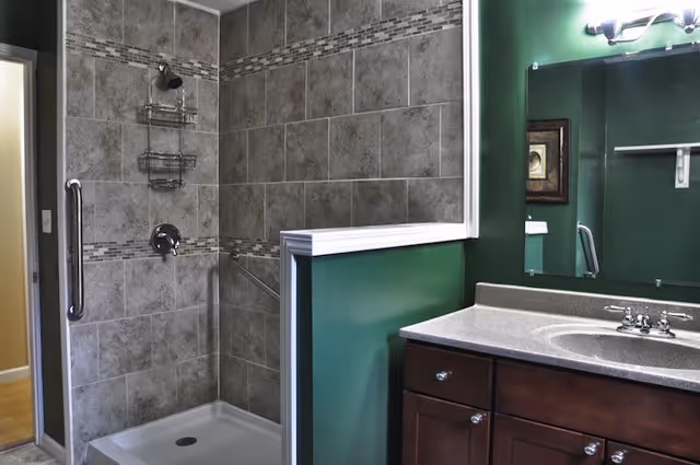 A bathroom featuring a tiled walk-in shower with a metal grab bar and a shower caddy. The walls are painted green with white trim. There is a dark wood vanity with a gray countertop and a sink, and a large mirror above it with a light fixture.