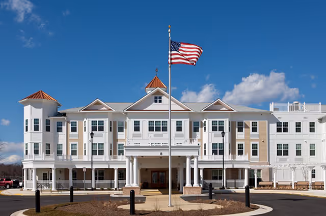 Front exterior view of a large senior living facility building with white and beige siding, multiple windows, and a central entrance with columns. An American flag flies on a flagpole in front of the building under a blue sky with some clouds.