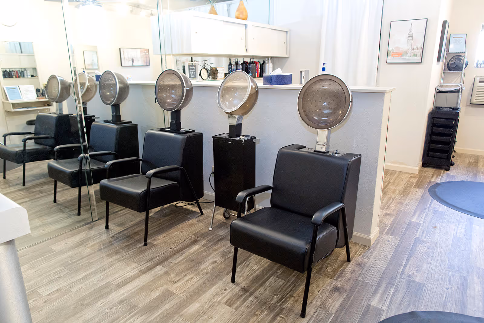 Interior of a hair salon area with three black salon chairs each equipped with a vintage hair dryer hood. The room has wood flooring, a large mirror on one wall, and shelves with hair products and decor items.