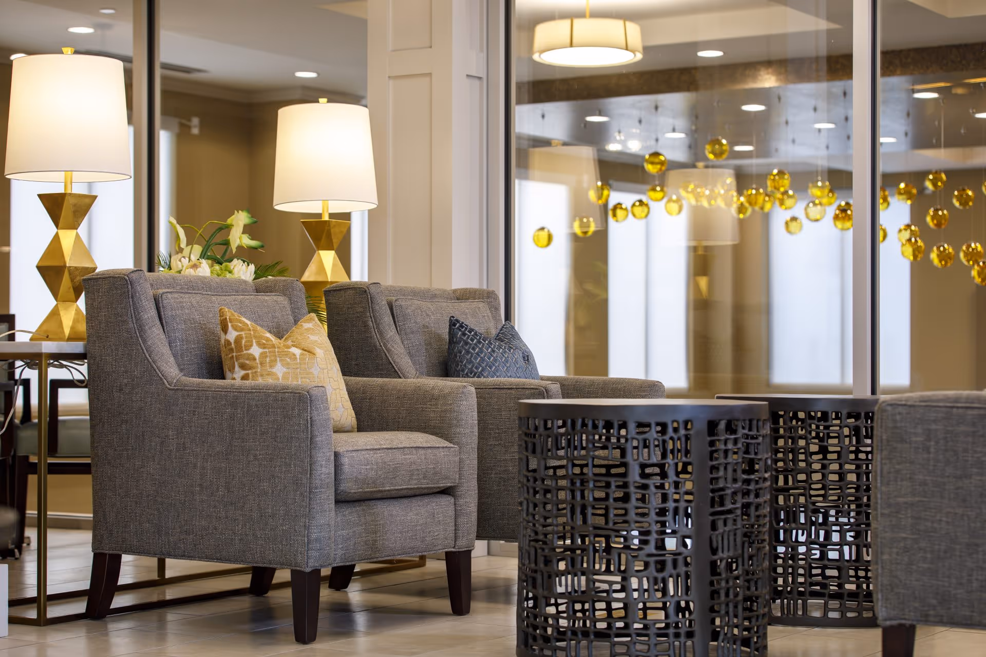 A modern lounge area with gray armchairs, patterned pillows, gold lamps, and decorative hanging golden orbs reflected in a glass wall.