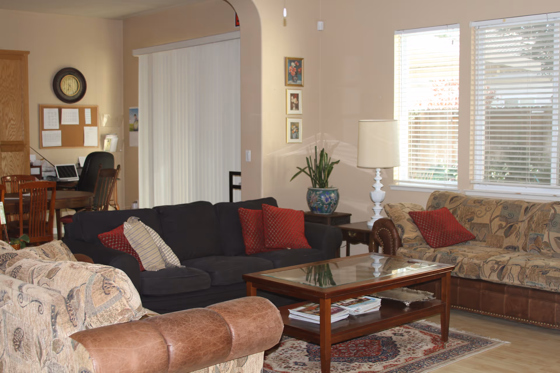 A cozy living room with two patterned beige sofas, one dark gray sofa with red and striped pillows, a wooden coffee table with magazines underneath, a side table with a white lamp and a potted plant, three framed floral pictures on the wall, and large windows with white blinds letting in natural light. In the background, there is a dining area with a wooden table, chairs, a clock, and a bulletin board on the wall.