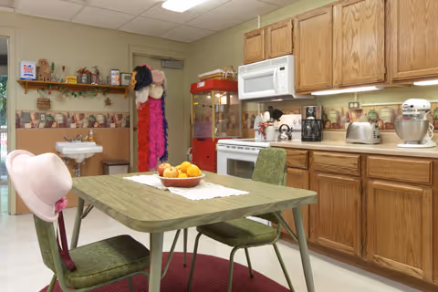 A kitchen area in a senior living facility with wooden cabinets, a microwave, stove, coffee maker, toaster, and mixer on the countertop. In the foreground, there is a green table with two green chairs, one with a pink hat hanging on its back. A bowl of fruit sits on a white doily on the table. In the background, there is a sink, a popcorn machine, and a rack with colorful hats and boas.
