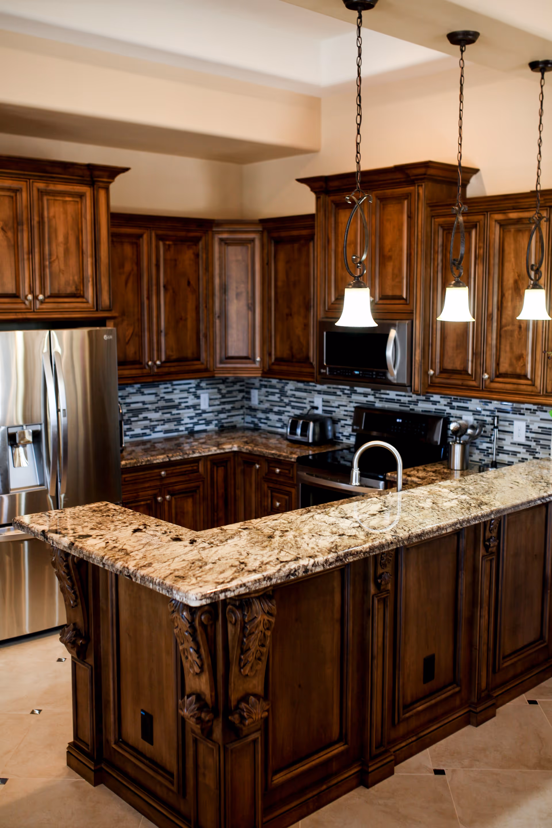 A kitchen with dark wooden cabinets, a granite countertop island with a built-in sink, stainless steel refrigerator, stove, microwave, and a toaster. The backsplash features a modern mosaic tile design, and three pendant lights hang above the island.