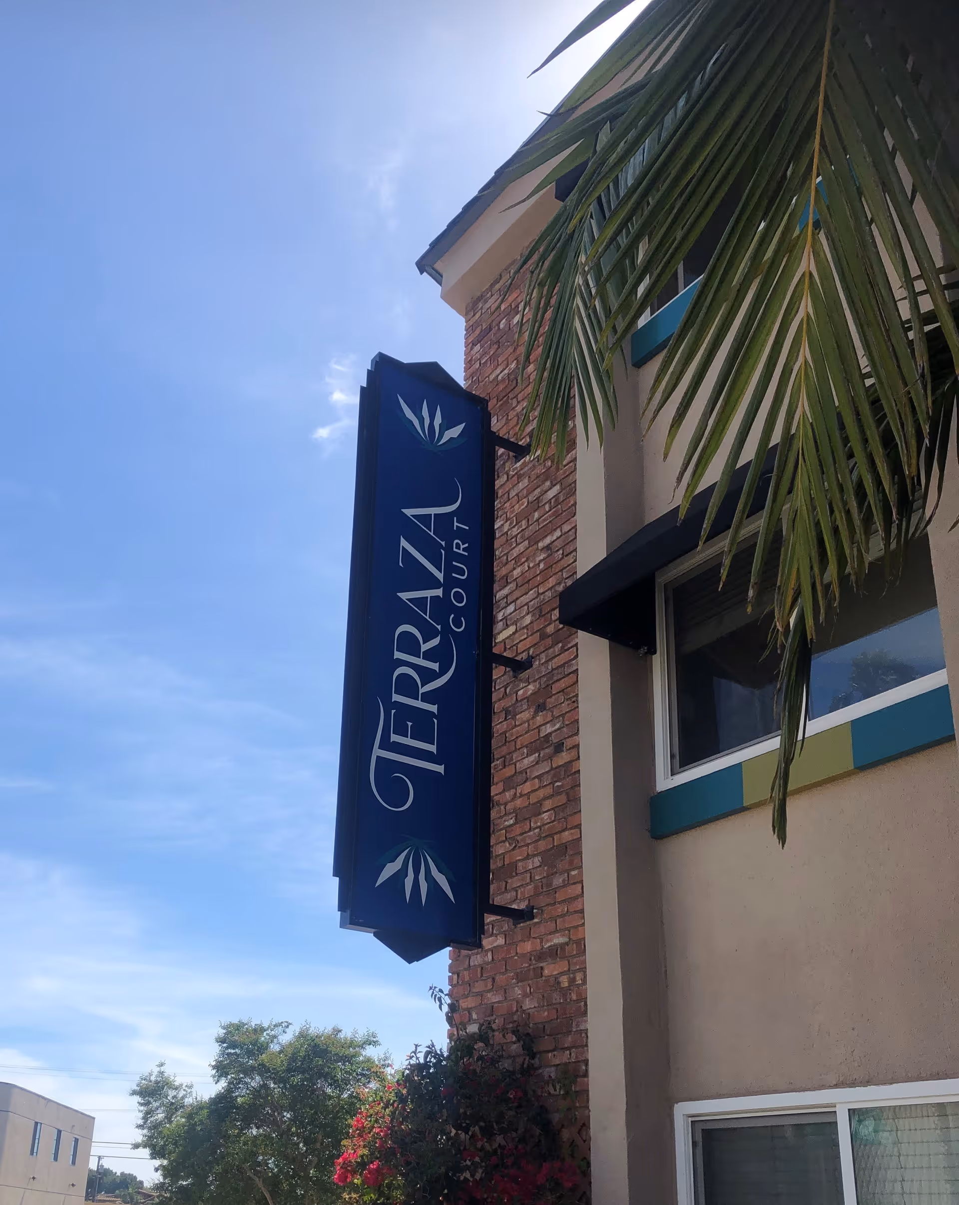 Exterior view of a building with a vertical sign that reads 'Terraza Court' mounted on a brick wall, with a palm tree branch partially visible in the foreground and a clear blue sky in the background.