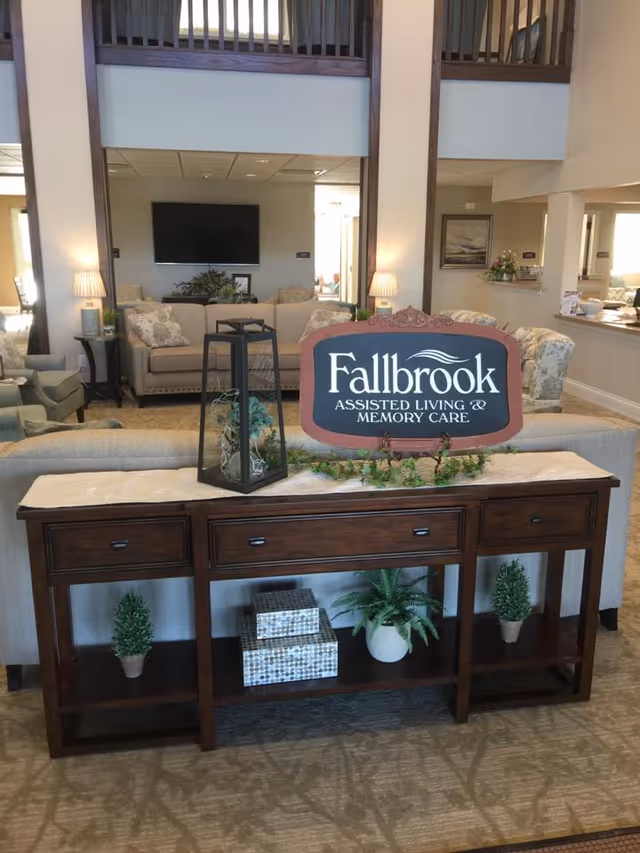 Interior lounge of an assisted living facility with a wooden console table displaying a 'Fallbrook Assisted Living & Memory Care' sign and seating area behind it.
