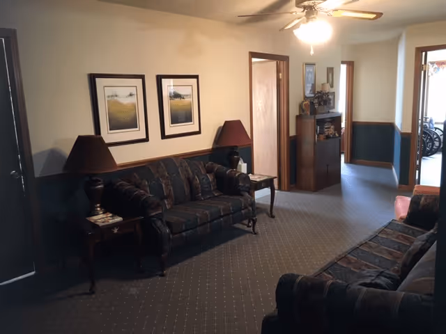 A living room area in Middleton Senior Apartments featuring two dark patterned sofas facing each other, two side tables with matching lamps, framed artwork on the wall, a ceiling fan with light, and a carpeted floor. The room has a doorway leading to another area with a wheelchair visible in the background.