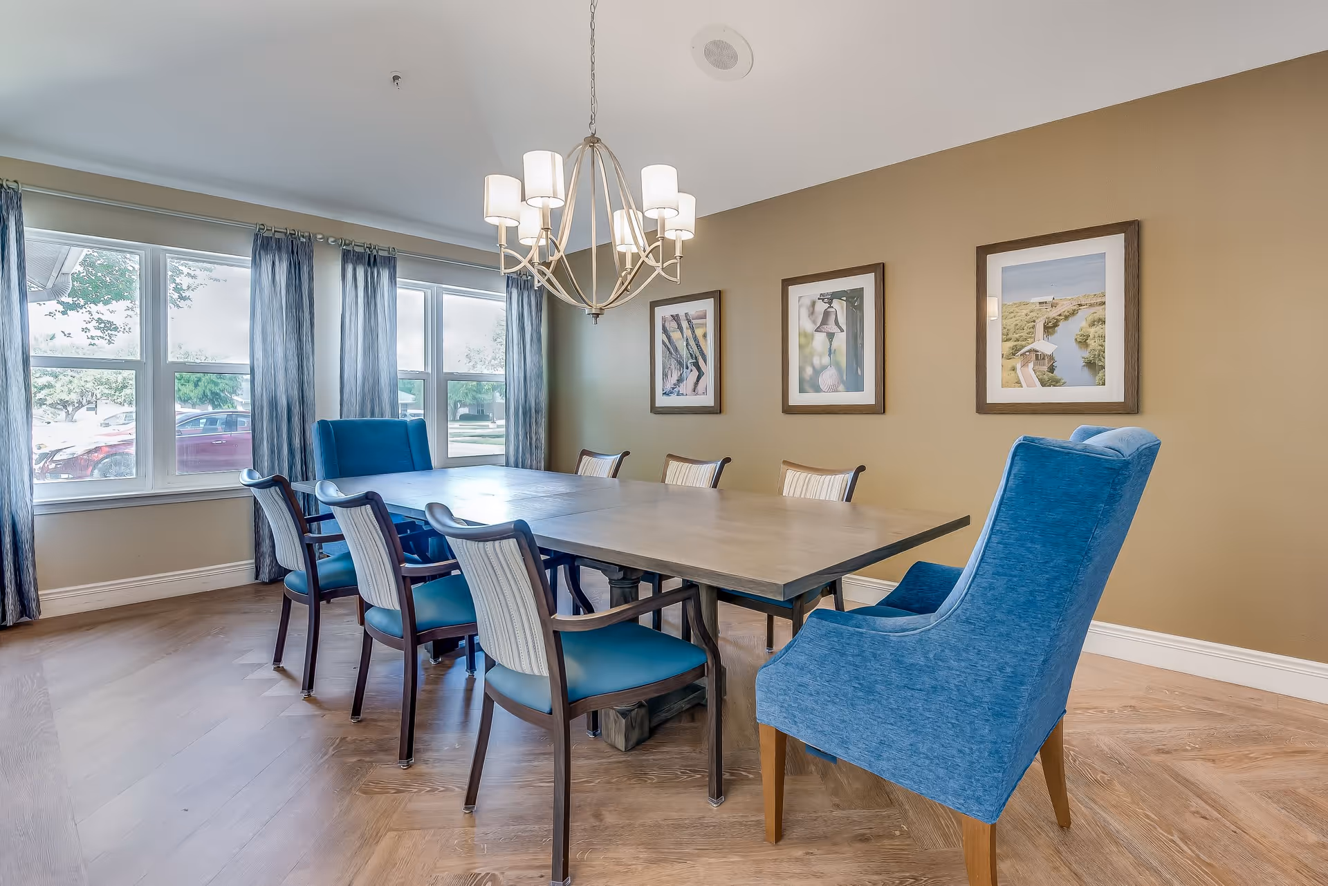 A dining room with a rectangular wooden table surrounded by eight chairs, including two blue upholstered armchairs at the ends and six wooden chairs with blue cushions. The room has large windows with blue curtains, a beige wall with three framed pictures, and a modern chandelier hanging above the table.