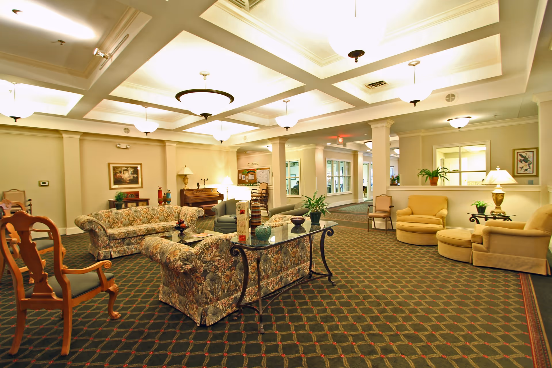 A spacious, well-lit living room area in a senior living facility with patterned carpet, floral upholstered sofas, beige armchairs, wooden chairs, a glass-top table with decorative items, potted plants, a piano, and framed artwork on the walls.