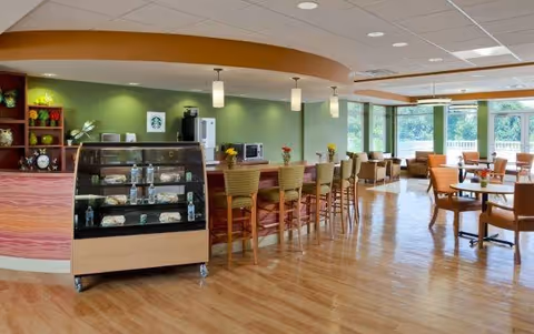 A bright and spacious dining area with wooden flooring, a display case with snacks and bottled water, a counter with high chairs, and several tables and chairs near large windows letting in natural light.