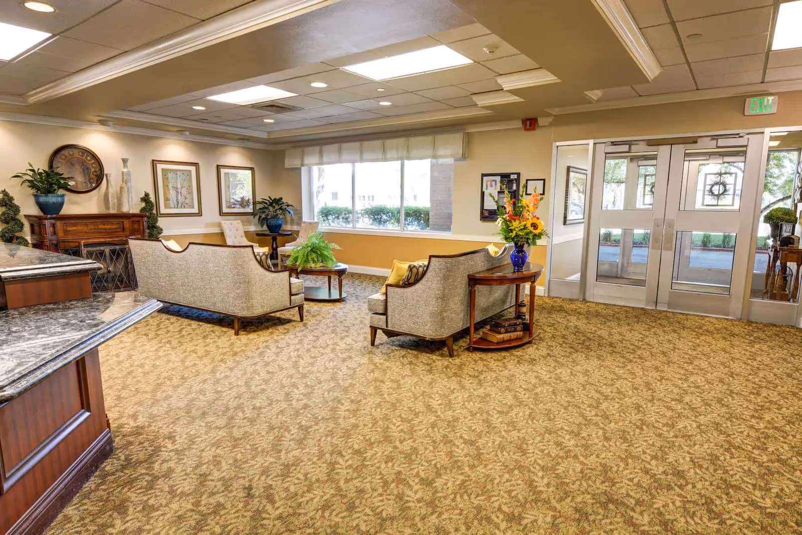 A spacious and well-lit senior living facility lounge area with beige patterned carpet, two upholstered sofas facing each other around a round wooden coffee table with a green plant. There are two beige chairs near a window with a view of bushes outside. The walls are decorated with framed artwork and a large clock. A wooden side table with a blue vase holding a colorful flower arrangement is next to one sofa. Double glass doors lead outside, and the ceiling has recessed lighting.