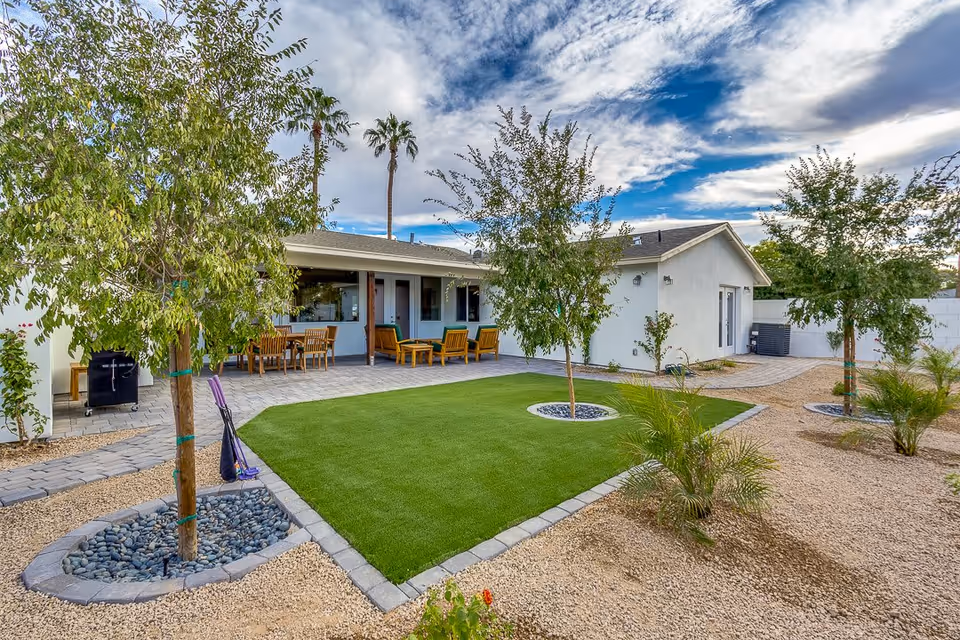 Outdoor patio area at Vista Living Arcadia featuring a green artificial turf lawn bordered by stone pavers, several young trees planted in circular stone beds, and a seating area with wooden chairs and tables under a covered porch. The sky is partly cloudy with patches of blue.