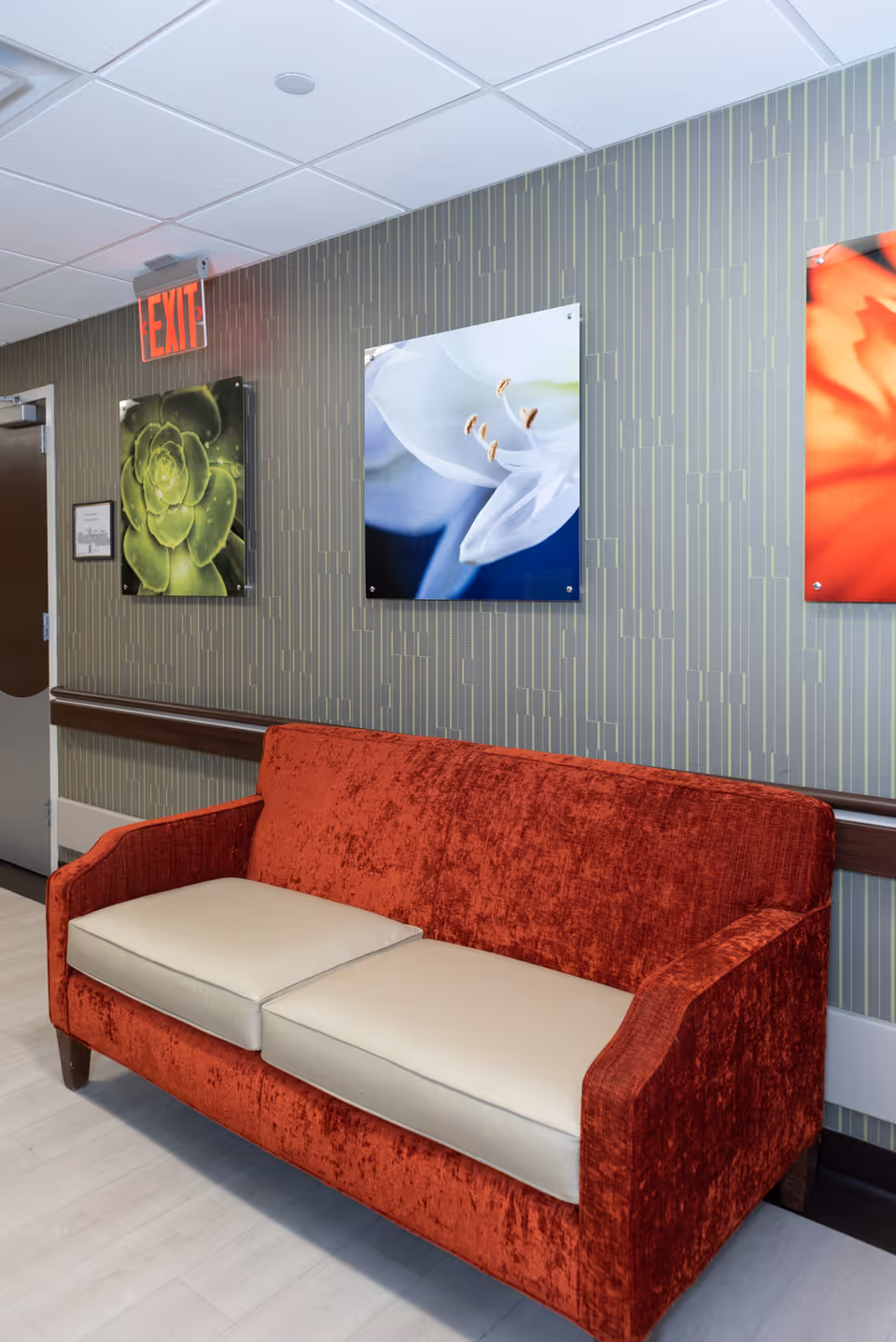 A red velvet sofa with beige cushions placed against a patterned wall in a hallway. Above the sofa, there are three framed floral photographs and an illuminated red EXIT sign near a door.