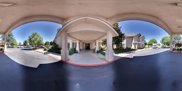 Covered entrance area of a senior living facility with a driveway and parking spaces on both sides. The building has beige walls and multiple windows, surrounded by green trees and shrubs under a clear blue sky.
