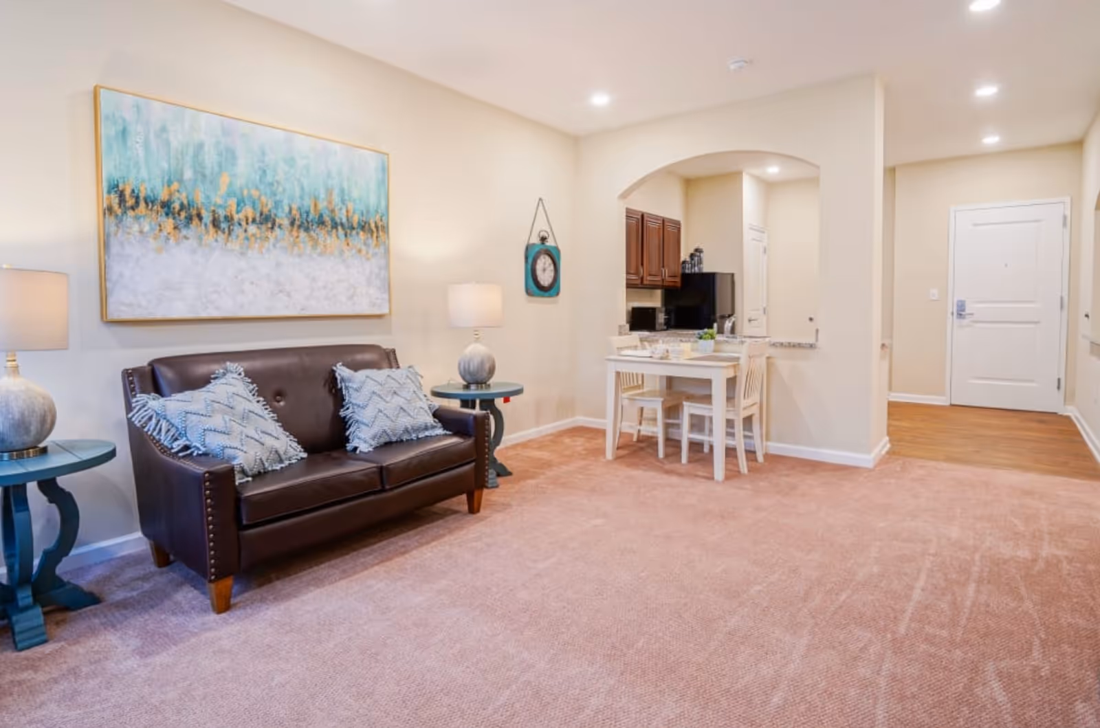 Well-lit living area with a brown leather sofa, side tables and wall art, a small dining table by a kitchenette, and the entry door visible.