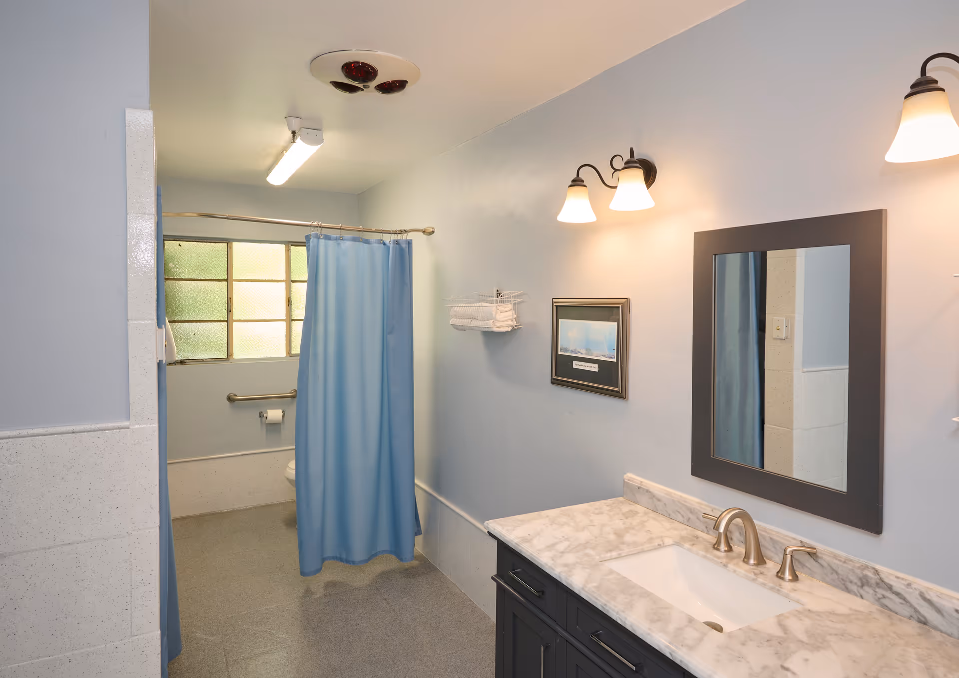 Accessible bathroom with a marble-topped vanity and mirror on the right and a shower with a blue curtain, grab bar, and toilet area on the left.