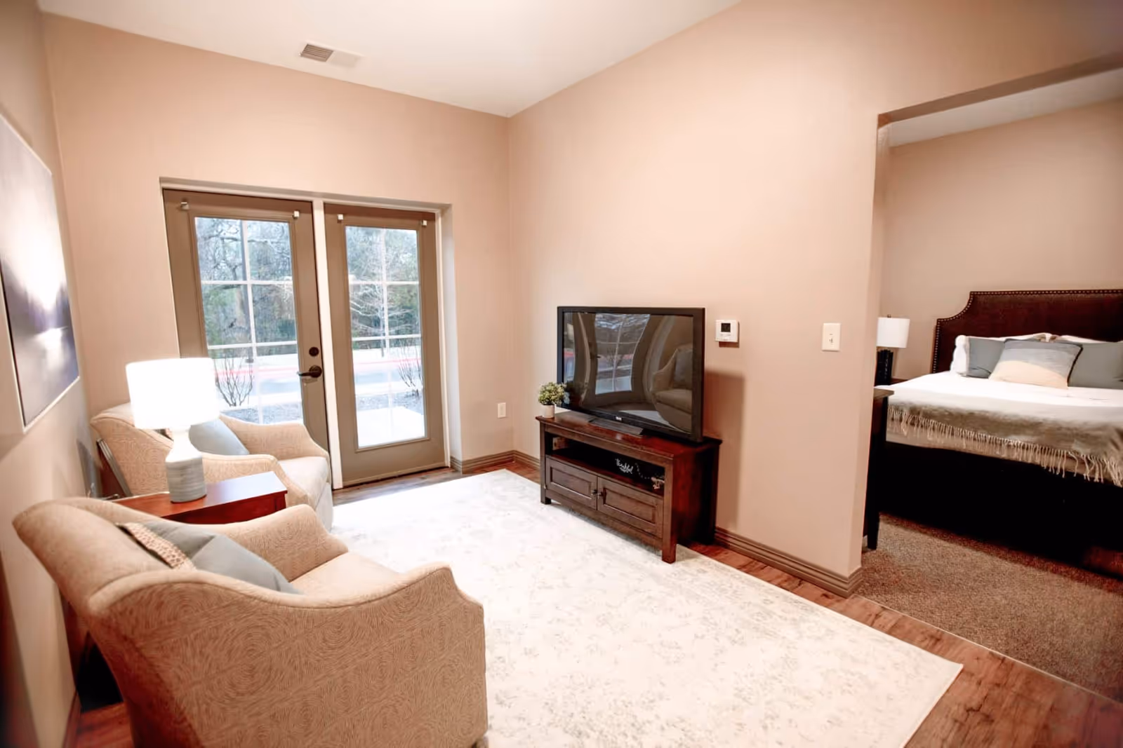 A cozy living room area with two beige armchairs, a small wooden side table with a lamp, a flat-screen TV on a wooden TV stand, and a large windowed door leading outside. Adjacent to the living room is a bedroom with a bed, pillows, and a bedside lamp visible through an open doorway.