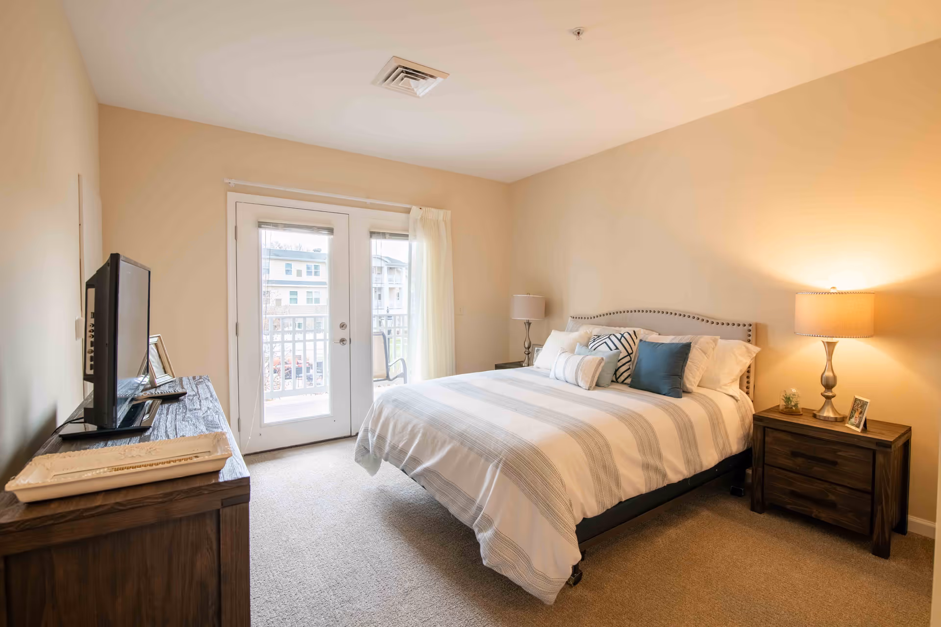Sunlit bedroom with a neatly made bed, nightstands and lamps, a TV on a dresser, and glass doors opening to a balcony.