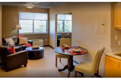 A cozy living area with a dark leather armchair, a round ottoman, a small round dining table with two chairs, and large windows letting in natural light. The room has beige walls and carpet, with a lamp on a side table next to the armchair.