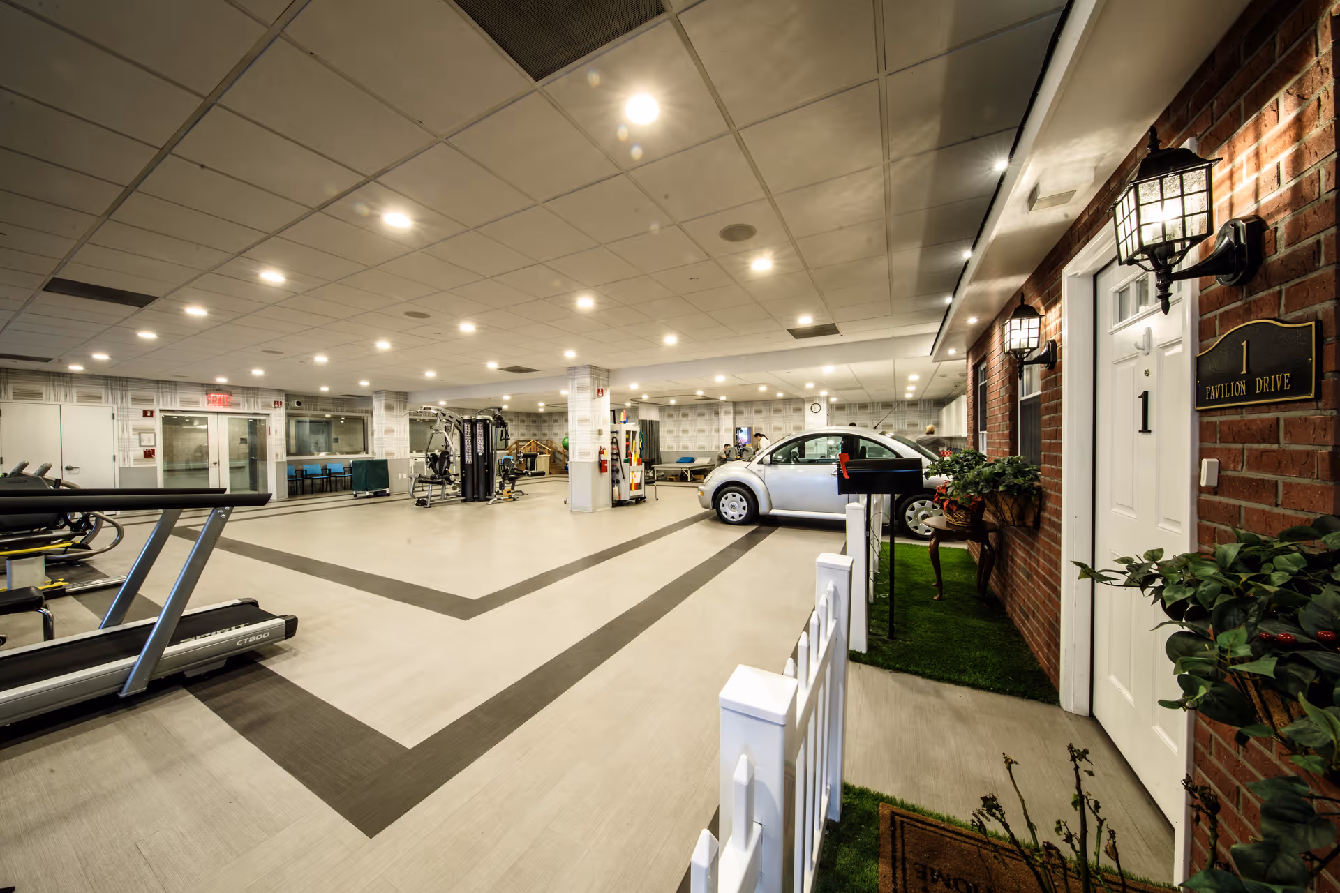 Indoor rehabilitation area featuring a treadmill, exercise equipment, a silver car, and a mock residential setup with a white door labeled '1' and a small white picket fence, under bright ceiling lights.