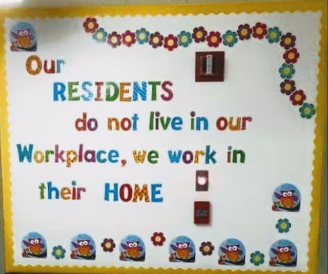 A bulletin board with a decorative yellow scalloped border and colorful flower cutouts. The board displays a message in multicolored letters that reads: 'Our RESIDENTS do not live in our Workplace, we work in their HOME.' There are small owl images along the bottom and corners of the board.
