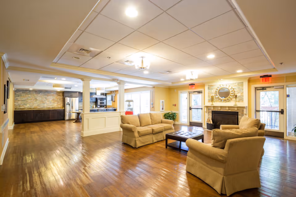 A spacious senior living facility common area with wooden floors, beige sofas, a coffee table, and a fireplace with a decorative mirror above it. The room is well-lit with ceiling lights and has large windows and glass doors leading outside. The kitchen area with stainless steel appliances is visible in the background.