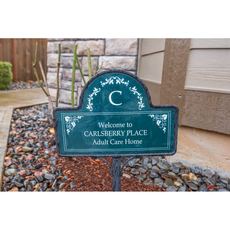 A green decorative sign in a garden bed with rocks and mulch, displaying the text 'Welcome to CARLSBERRY PLACE Adult Care Home' in front of a building with stone and wood siding.