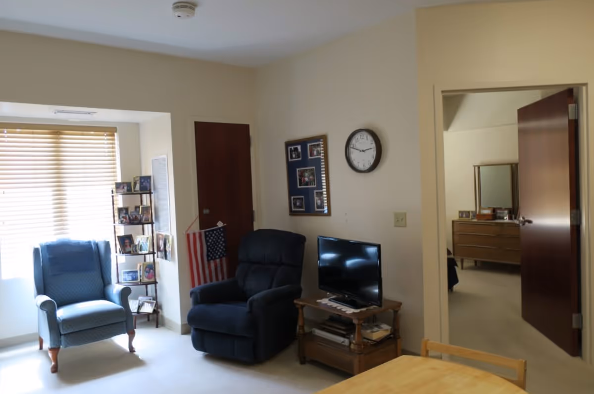 A cozy living room area with two armchairs, one blue and one dark blue, positioned near a window with blinds. A small wooden table holds a flat-screen TV and some books. On the wall above the TV is a clock and a bulletin board with photos. An American flag is displayed next to the dark blue chair. An open doorway leads to a bedroom with a dresser and mirror visible.